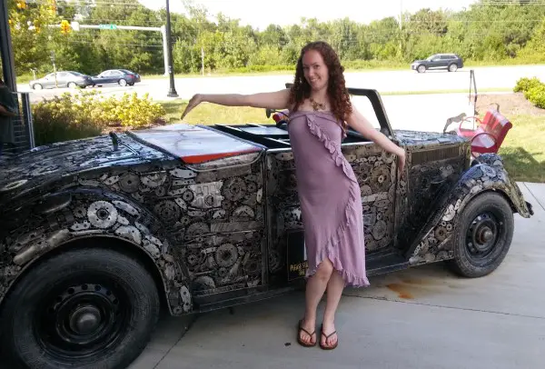 woman in purple dress posing in front of scrap metal car sculpture