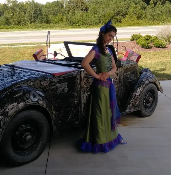 woman wearing peacock dress standing in front of scrap metal car sculpture