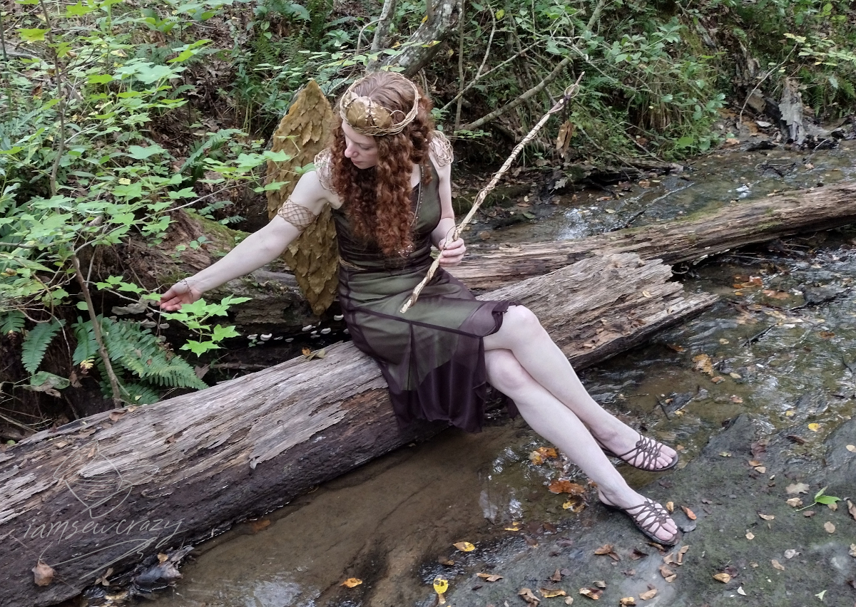 red haired woman wearing homemade forest fairy costume sitting on fallen log in creek